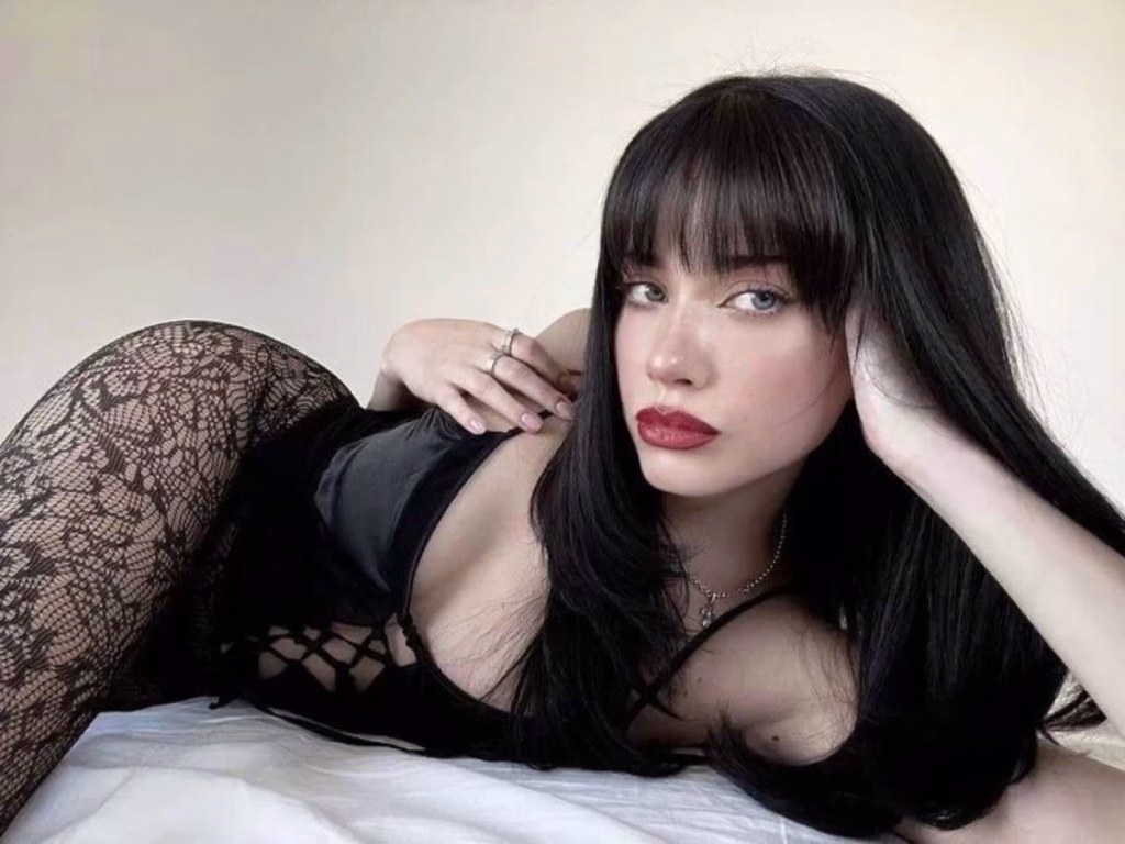 Woman with long black hair in black lace tights and a black outfit lying on a bed and looking confidently at the camera.